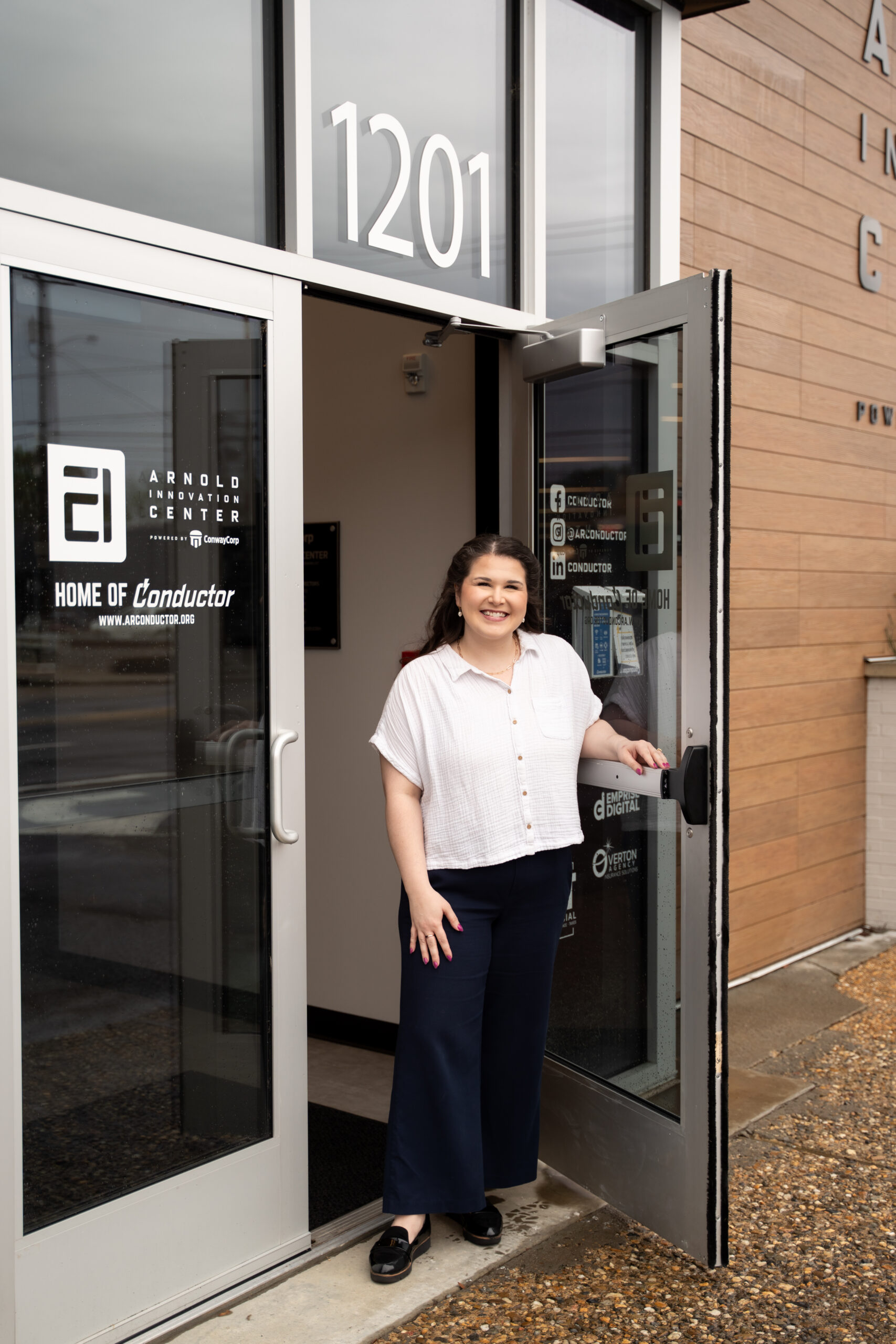 Conductor-38 (1) A woman smiling stands at the open glass door of a building with 1201 above the entrance and Arnold Innovation Center signage on the glass. She is wearing a white blouse and dark pants.