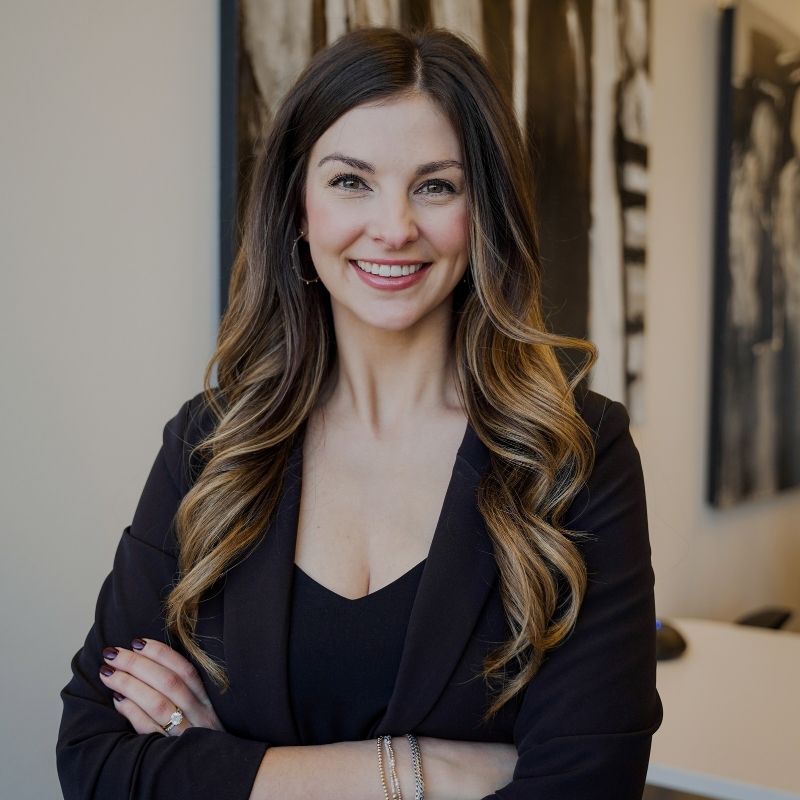Conductor Headshot (1) A woman with long wavy brown hair, wearing a black blazer and top, stands smiling with arms crossed in a modern office setting with abstract artwork in the background.