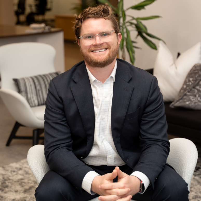 A man with glasses and a beard, wearing a dark suit and white shirt, sits smiling in a modern office with stylish chairs, a plant, and decorative pillows in the background.