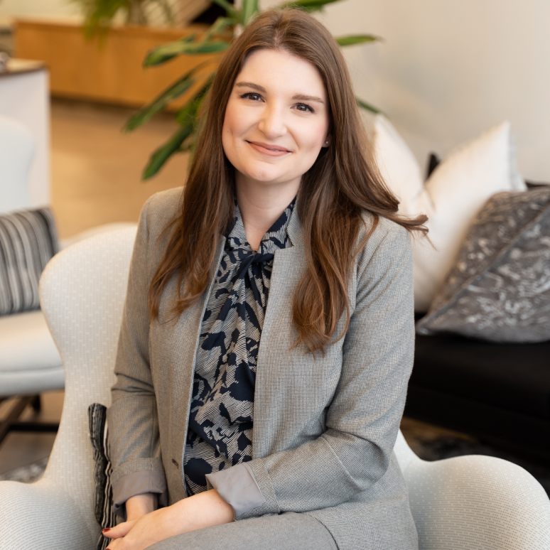 A woman with long brown hair, wearing a grey suit and patterned blouse, sits smiling in a modern, comfortable office setting with plants and cushions in the background.