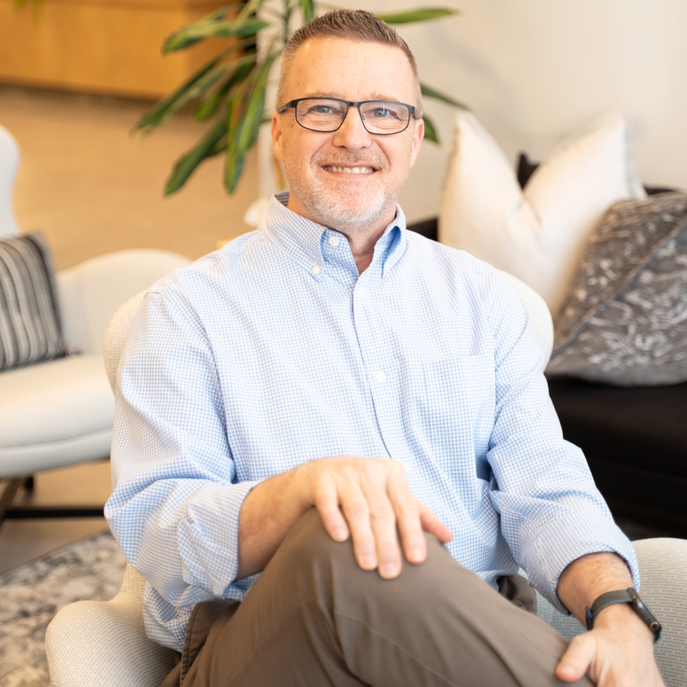 A smiling man with glasses, short hair, and a light blue checkered shirt sits with his legs crossed in a modern, well-lit living room with neutral-colored decor and cushions.