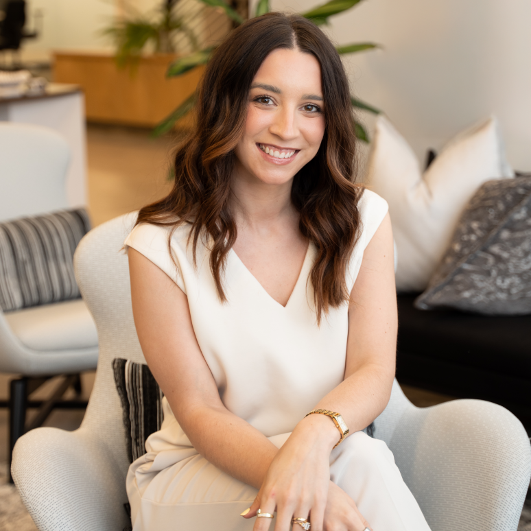 A woman with long, wavy brown hair and a white outfit sits smiling in a modern, stylish office space with neutral decor and patterned pillows.