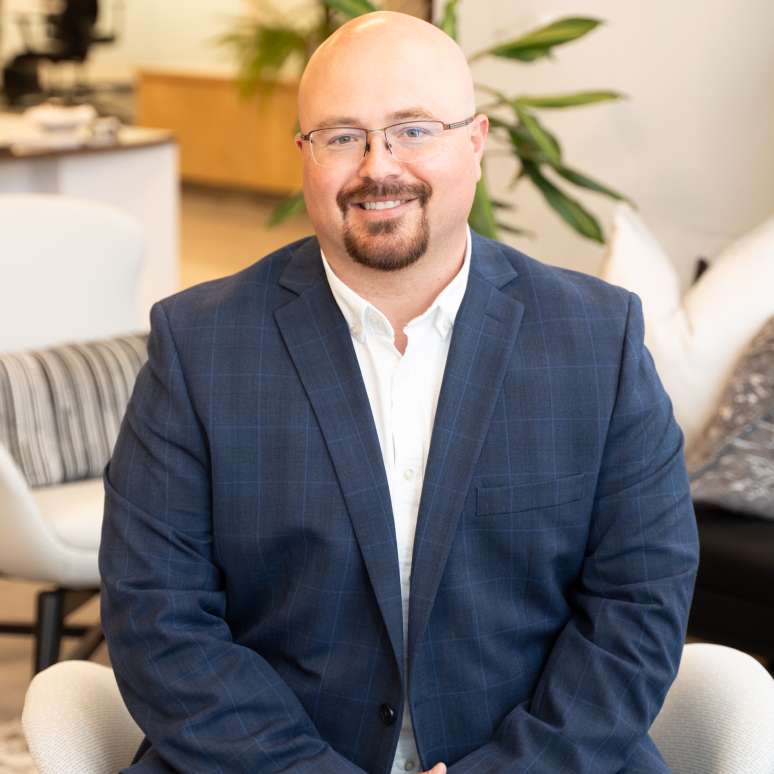 A bald man with glasses and a short beard, wearing a blue checked suit jacket and white shirt, sits smiling in a modern office setting with plants and neutral-colored furniture in the background.
