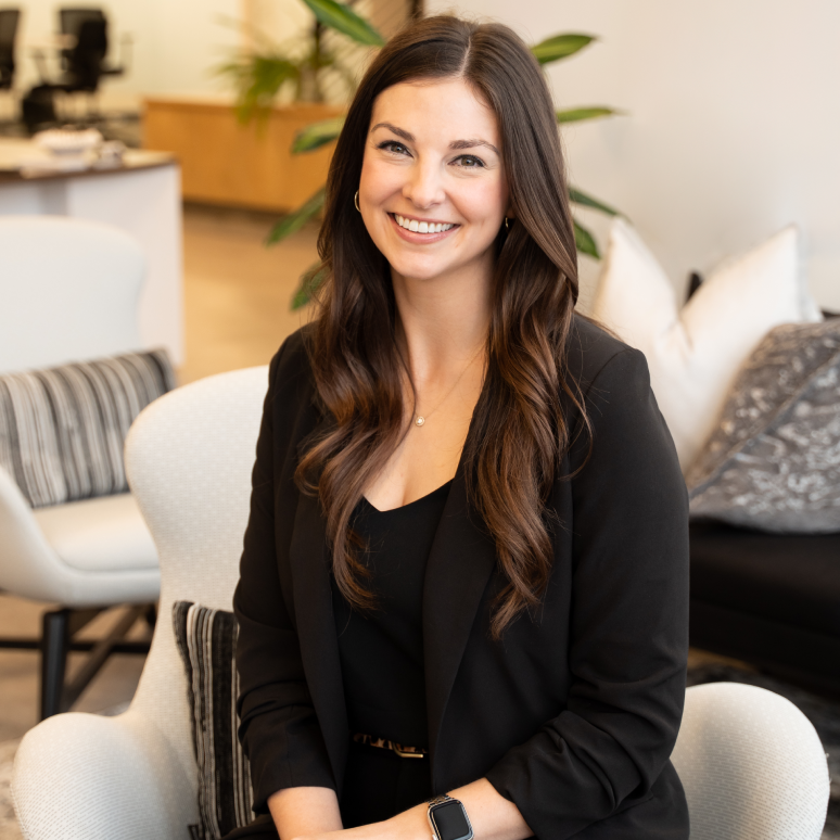 A woman with long brown hair, wearing a black blazer and black top, sits smiling on a white chair in a modern office setting with plants and neutral-toned decor in the background.