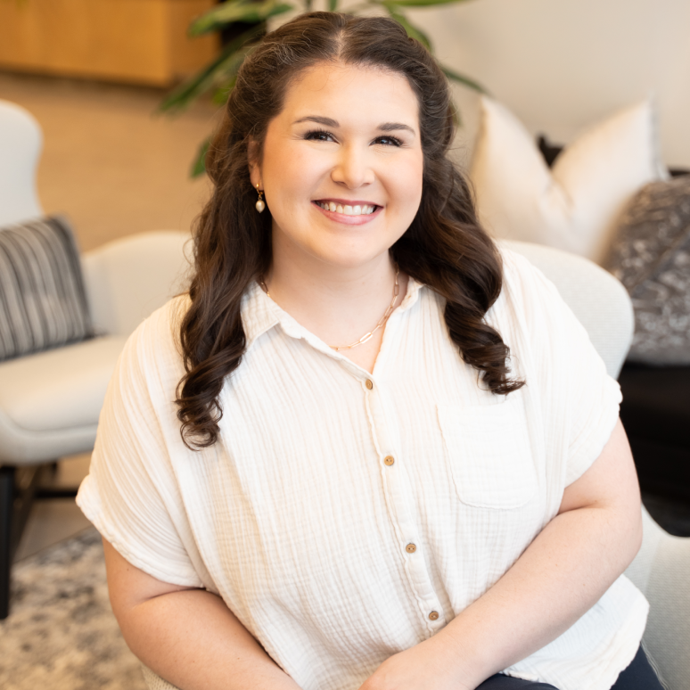 A woman with long, wavy brown hair sits smiling in a bright, modern room. She wears a short-sleeve white button-up shirt and gold earrings. Cushioned chairs, pillows, and a plant are visible in the background.