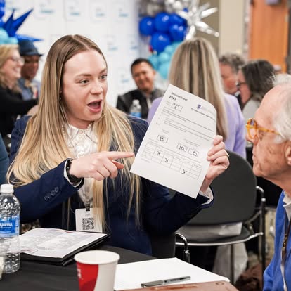 A woman with long blond hair excitedly points to a form she is holding up while sitting at a table in a busy, festive room. People are gathered in the background, and balloons are visible.