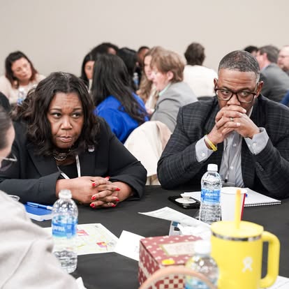 A group of people sit around a table engaged in discussion at a meeting or conference. Two individuals in the foreground appear thoughtful, with bottled water and papers on the table.