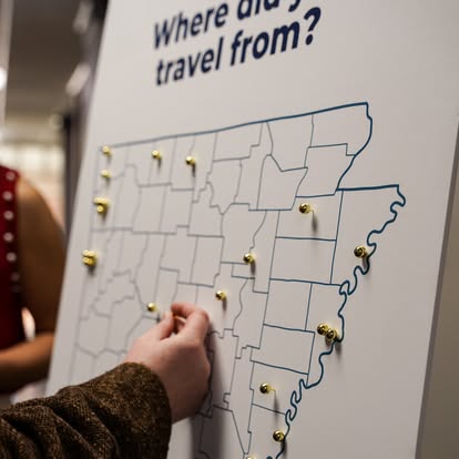 A person places a pin on a large map with outlined regions, under the text Where did you travel from? Multiple pins mark various locations on the map. Another person stands nearby.