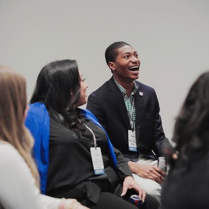 A group of four people sit together, smiling and talking. One man in a dark blazer and checked shirt is laughing. All are wearing name badges and appear to be at a professional event or conference.