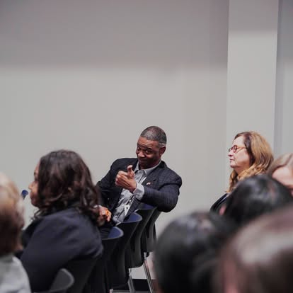 A man sitting among a group of people in a conference room gestures with a thumbs up, smiling. Others seated nearby listen attentively, and the setting appears to be a formal event or meeting.