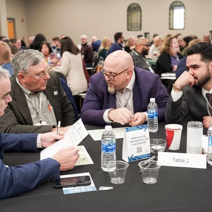 Four men in business attire sit at a round table in a crowded conference room, engaged in discussion with papers, water bottles, phones, and name cards in front of them. A “Table 2” sign is visible on the table.