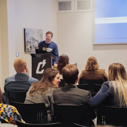 A group of people seated in a conference room listens to a speaker standing at a podium, with a projection screen and a picture on the wall in the background.