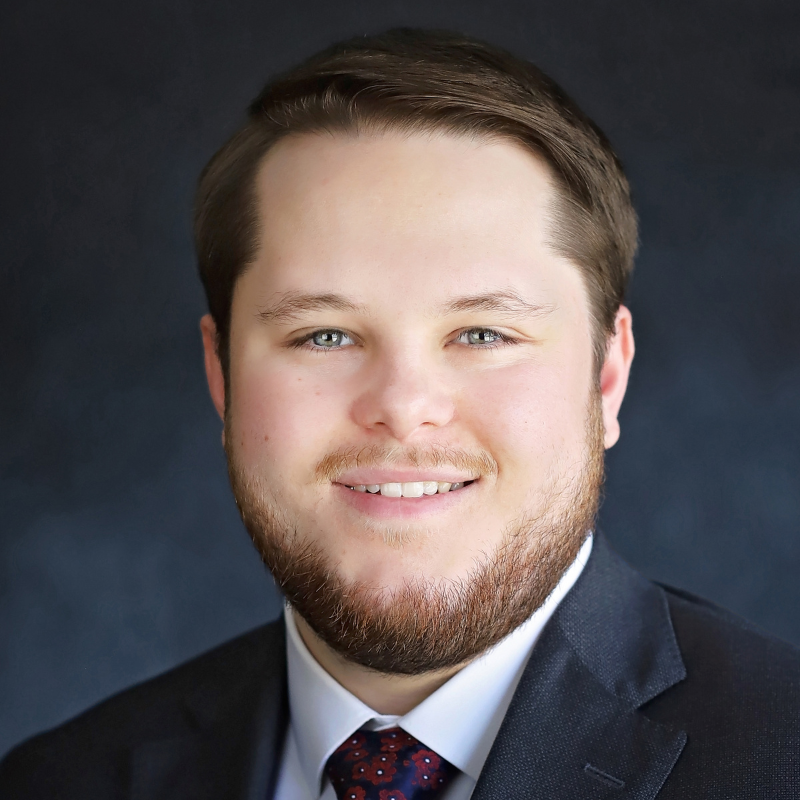 A man with short brown hair, a trimmed beard, and light-colored eyes, wearing a dark suit, white shirt, and patterned tie, smiling against a dark, blurred background.