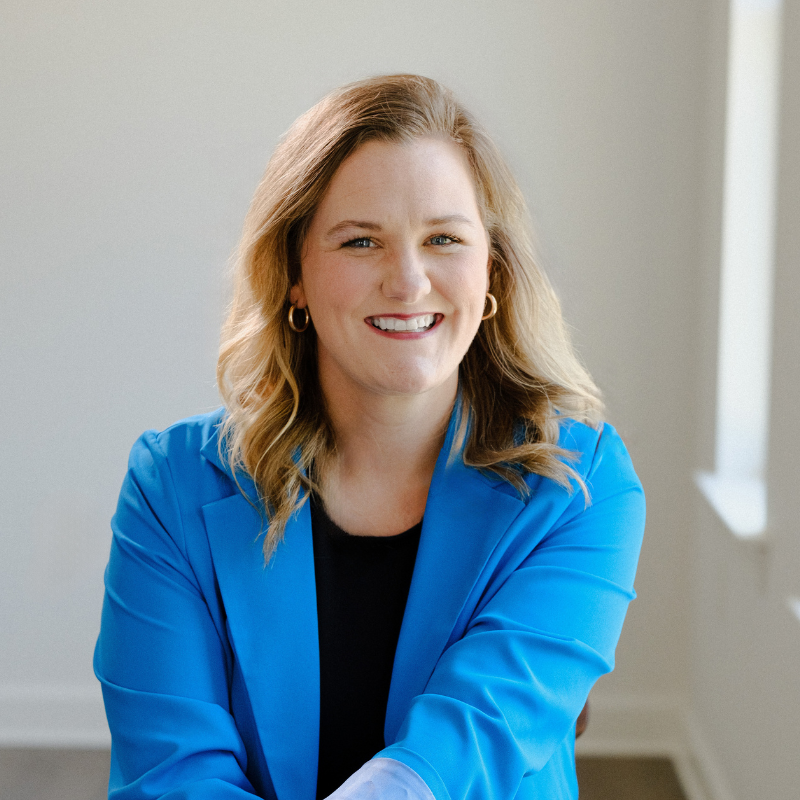 A woman with wavy blonde hair, wearing a bright blue blazer and gold hoop earrings, smiles while sitting indoors in a brightly lit room.