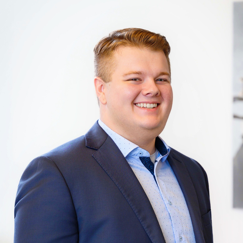 A young man with short, light brown hair wearing a dark suit jacket and a light blue dress shirt smiles while standing in front of a plain white background.