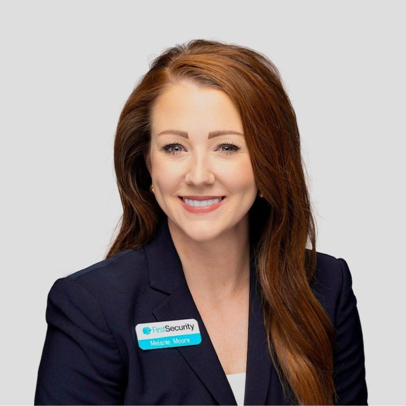 A woman with long auburn hair wearing a dark blazer and a name tag, smiling against a plain white background.