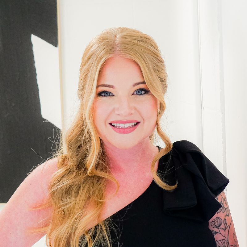 Smiling woman with long, wavy blonde hair wearing a black one-shoulder top, standing indoors in front of a white wall with a black and white abstract painting.