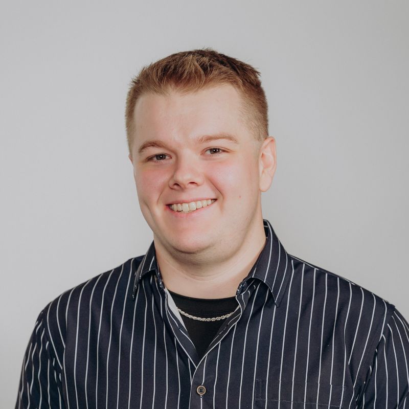 A young man with short light brown hair smiles at the camera. He is wearing a black shirt with white vertical stripes over a black t-shirt and a thin silver chain. The background is plain and light gray.