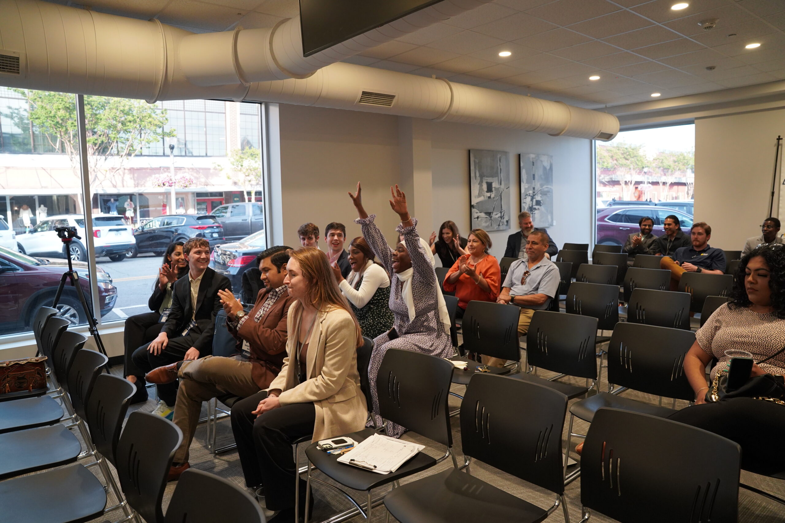 People seated in a conference room, some raising their hands to participate. Several empty chairs are visible, and large windows show cars and trees outside. A notebook and phone rest on a chair in the front row.