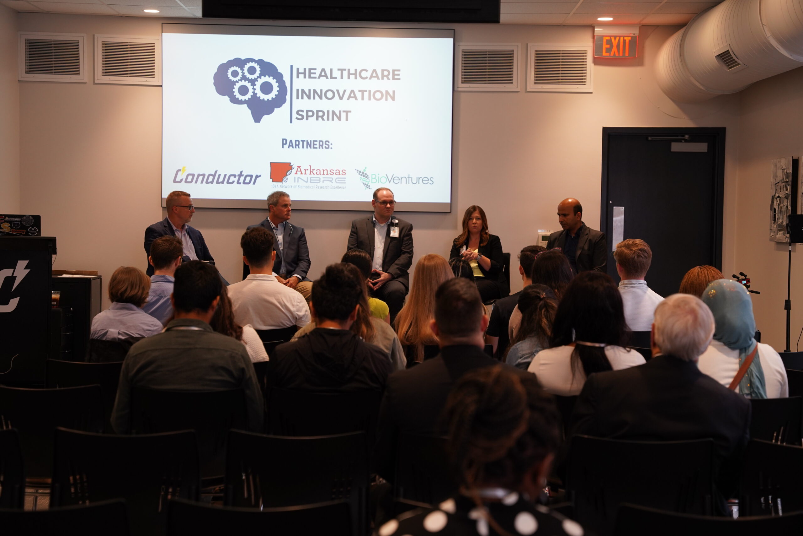 A panel of five professionals discusses onstage at a Healthcare Innovation Sprint event, facing an audience. A presentation slide with logos and partner names is projected behind them.