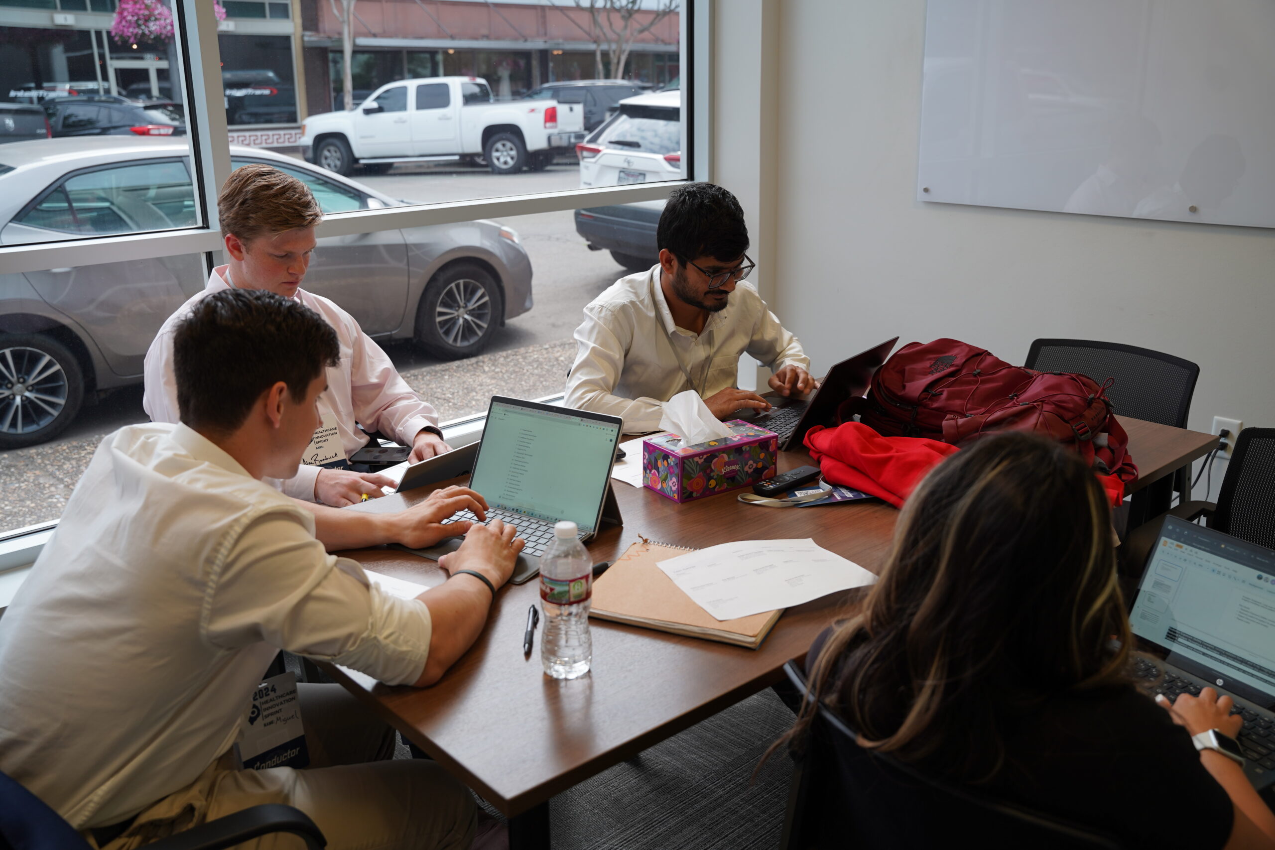 Four people sit around a table working on laptops in a bright meeting room with large windows. Papers, a water bottle, tissue box, and a red bag are on the table. Cars are visible outside the window.