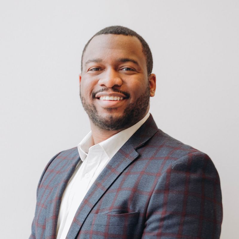 A man with short hair and a beard, wearing a checked blazer and white shirt, smiles while standing against a plain light background.