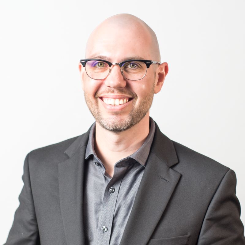 A smiling man with glasses and a shaved head, wearing a dark suit jacket over a gray button-up shirt, stands against a plain white background.