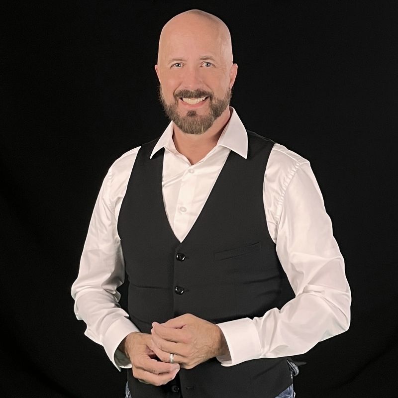 A bald man with a trimmed beard, wearing a white dress shirt and a black vest, stands smiling against a plain black background with his hands loosely clasped in front of him.