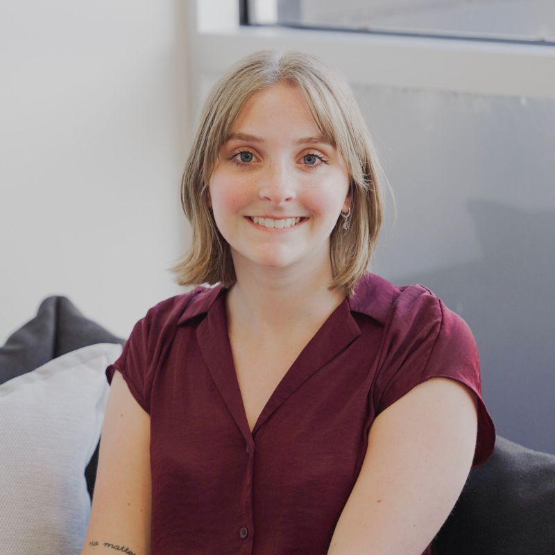 A young person with short, light brown hair and a burgundy shirt sits on a couch, smiling at the camera. There’s a window and a light-colored wall in the background.