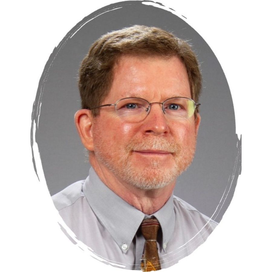 Portrait of an older man with short brown hair, glasses, and a trimmed beard, wearing a light-colored shirt and patterned tie, set against a gray background with a white oval frame.